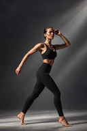 A woman in a black MOXII MOUVE sports bra and high-waisted MOXII MOUVE black leggings poses dynamically against a dark gray studio background.
