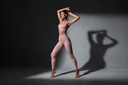 A woman modeling a MOXII MOUVE mauve pink activewear set consisting of a sports bra and high-waisted leggings, posed dynamically against a dark studio background with dramatic lighting.