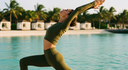 A woman in MOXII MOUVE olive green activewear set poses in a yoga pose by a tropical swimming pool with palm trees and resort cabanas in the background.